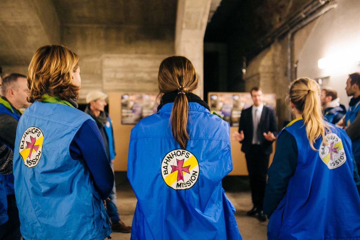Frauen in blauen Westen mit dem Logo der Bahnhofsmission verfolgen einen Vortrag eines Mannes