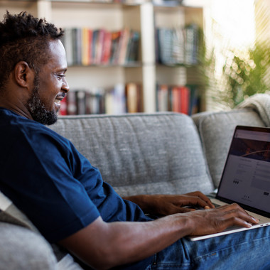 Mann sitzt mit ausgestreckten Beinen auf einem Sofa und arbeitet am Laptop, im Hintergrund Bücher und Pflanzen