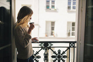 Frau mit Kaffeetasse steht an ihrem französischen Balkon und schaut aus der Wohnung