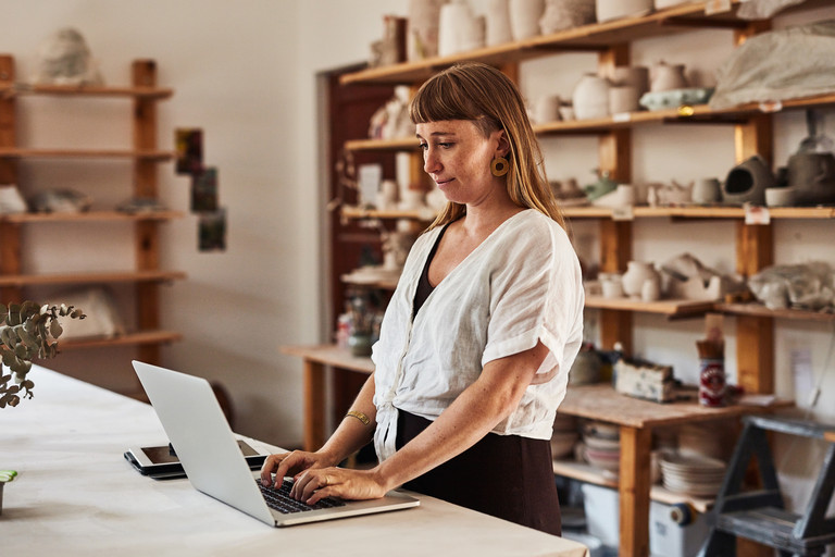 Junge Frau im Keramikstudio plant am Laptop, ihr Online-Business zu starten. Regale voller handgemachter Keramik im Hintergrund.