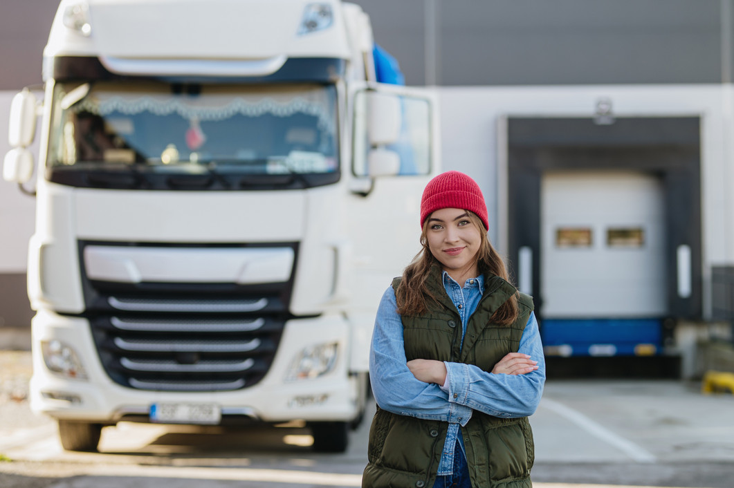 Frau mit einer roten Mütze steht mit verschränkten Armen vor einem großen LKW und lächelt