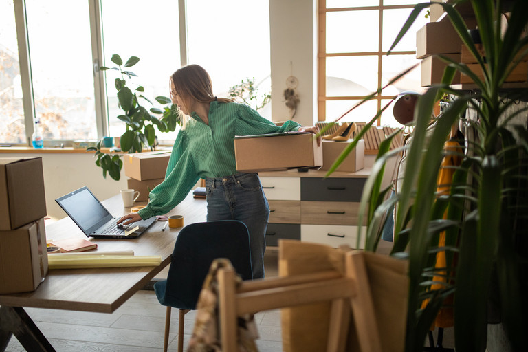 Frau mit Karton steht an einem Tisch mit Laptop und Paketen in einem hellen Raum mit Pflanzen und Verpackungsmaterial.