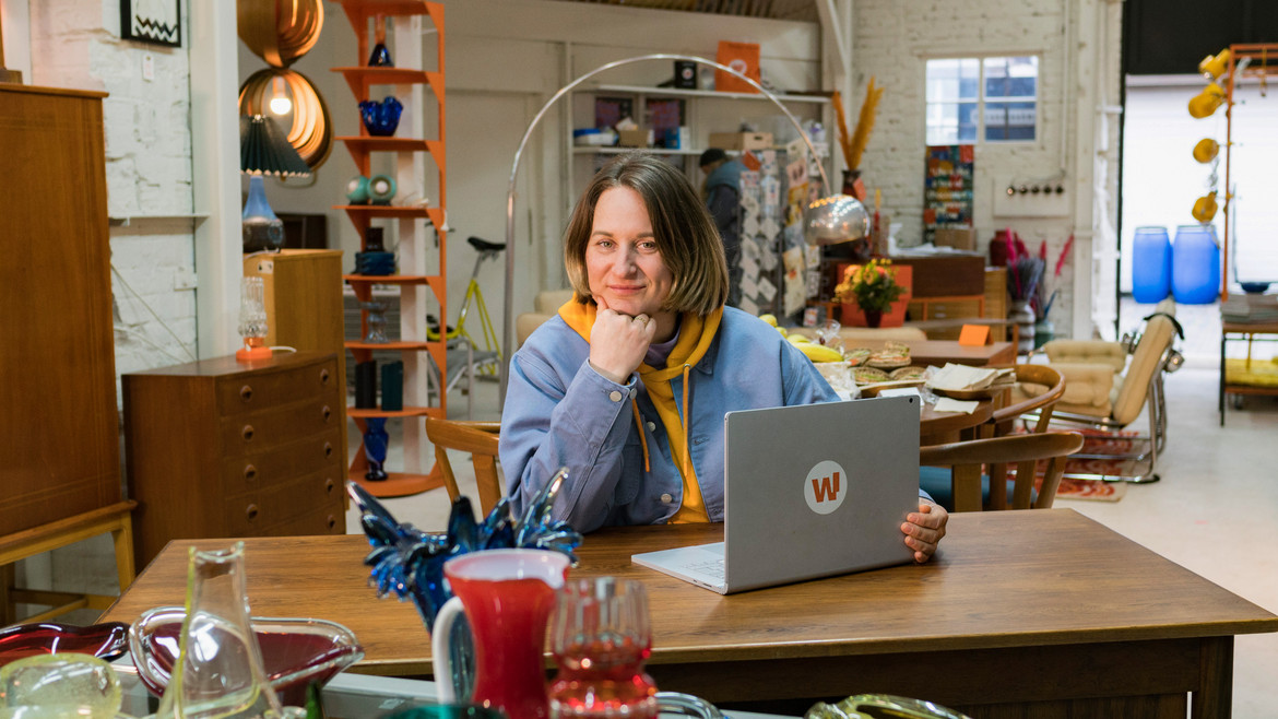 Lydia Wunderwald stützt sich mit dem Ellbogen auf einem Tisch ab und hat ihren Laptop vor sich, im Hintergrund befinden sich Lichter und Möbel