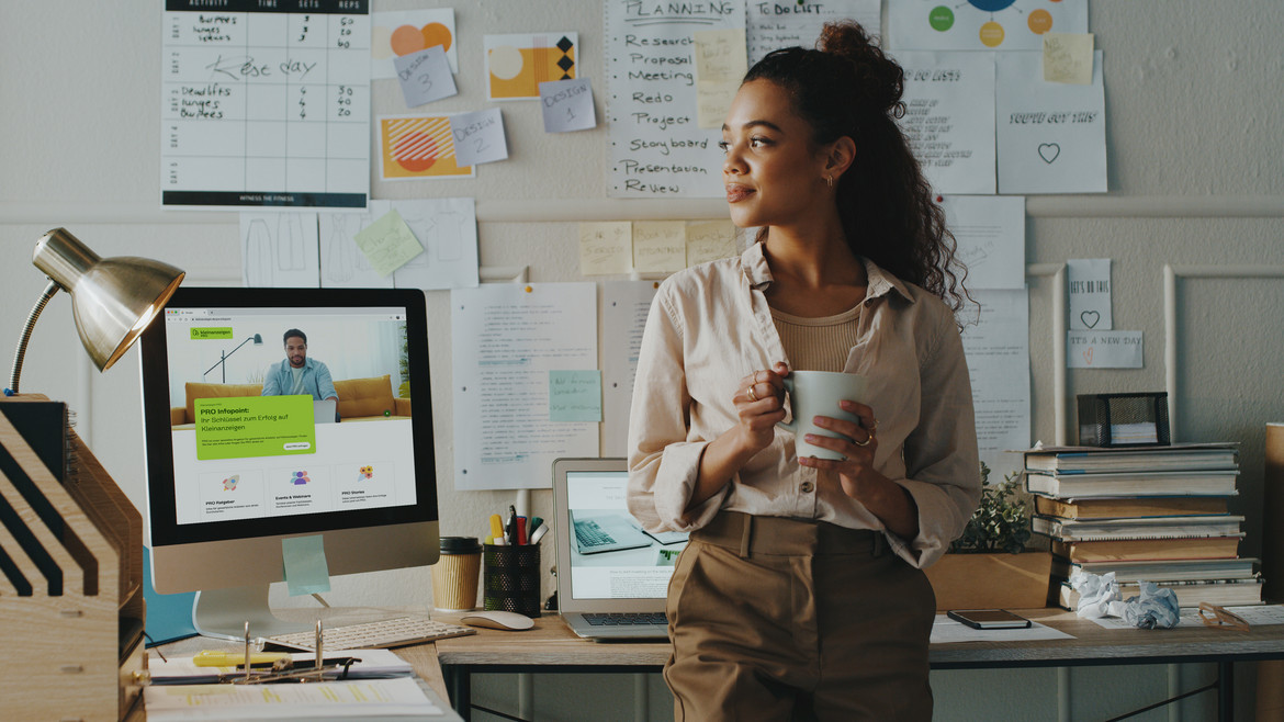 Frau mit Kaffeetasse steht im Büro mit Notizen an der Wand, Computer zeigt die Benutzeroberfläche von Kleinanzeigen PRO.