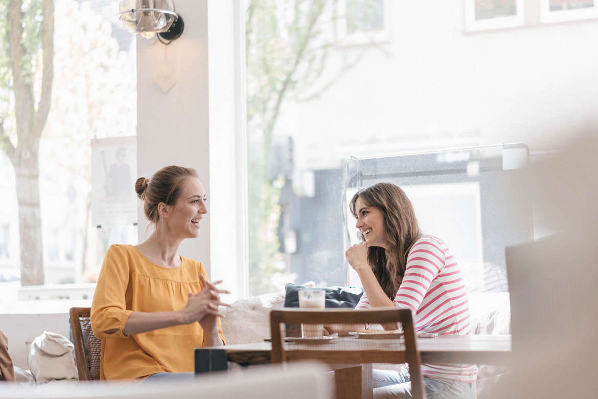 Zwei Junge Frauen sitzen an einem Tisch und unterhalten sich gut gelaunt