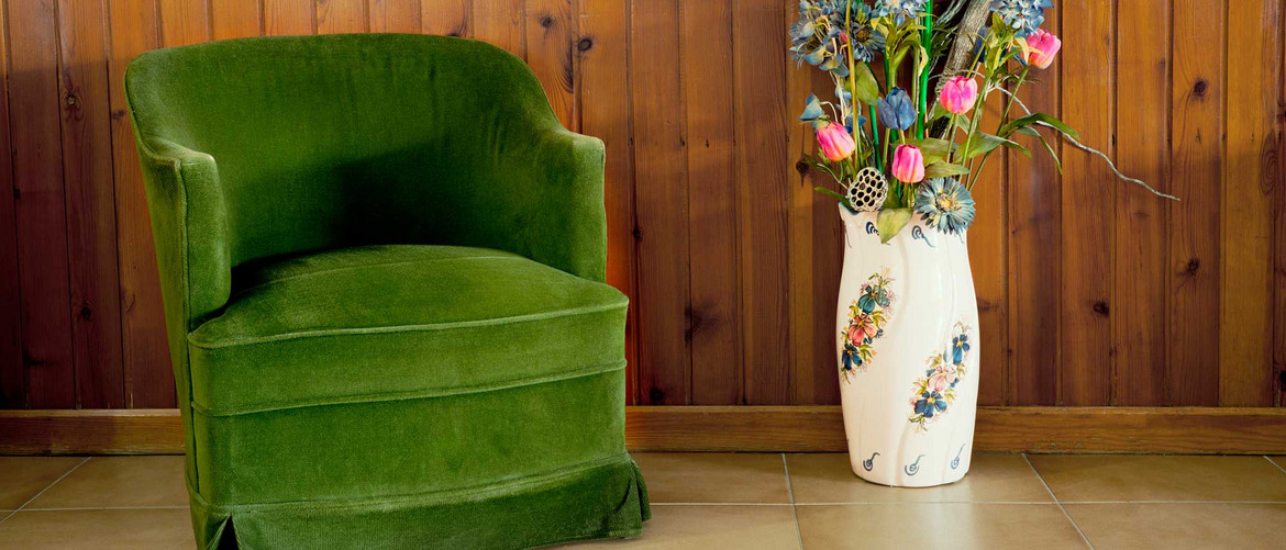 Grüner Stoffsessel im Retro-Design neben einer großen Blumenvase vor einer Holzwand