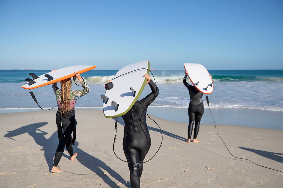 Drei Personen tragen Surfboards auf dem Kopf und gehen auf dem Strand in Richtung Wasser