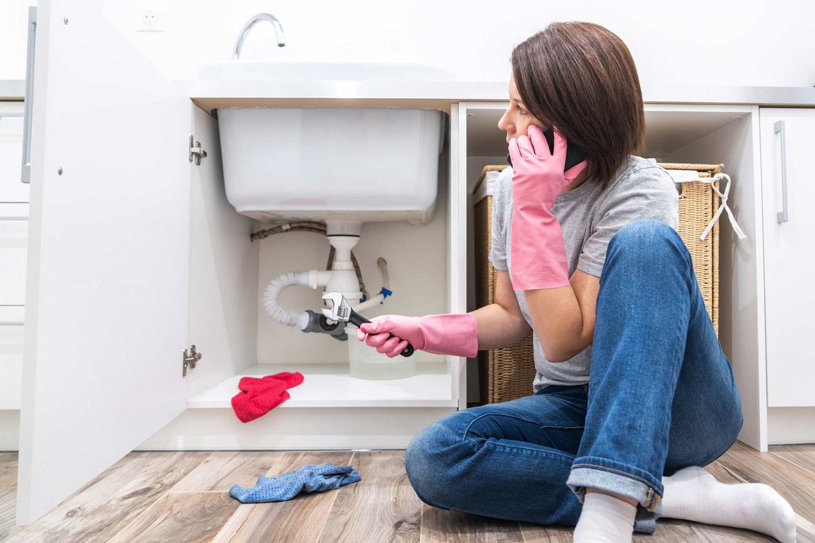 Frau sitzt mit Handschuhen und Zange vor Waschbecken und telefoniert mit Handwerker