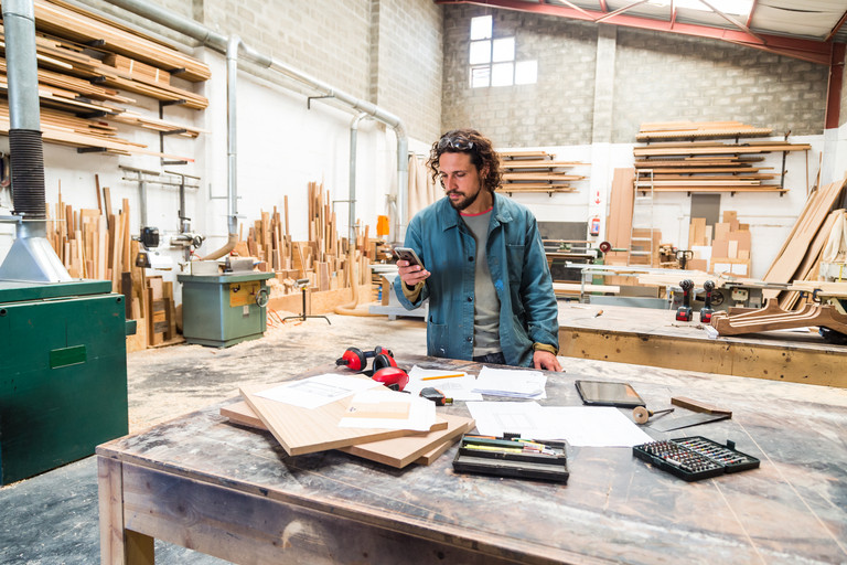 Mann mit lockigem Haar in einer Werkstatt steht am Tisch mit Holzplatten und Werkzeugen und schaut auf sein Smartphone.