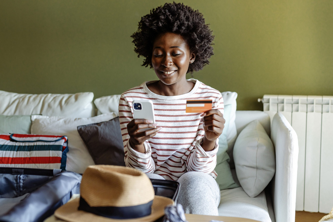 Frau sitzt auf einem Sofa, schaut in ihr Handy und hält eine Kreditkarte in der Hand.